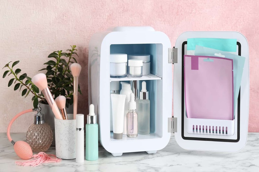 Tủ lạnh mini chứa mỹ phẩm, bên cạnh là cọ trang điểm và bình thủy tinh, nền hồng và cây trang trí.