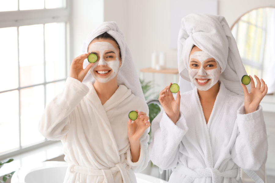 Hai người mặc áo choàng trắng, đắp mặt nạ, cầm lát dưa chuột, đứng cạnh cửa sổ trong phòng tắm.
