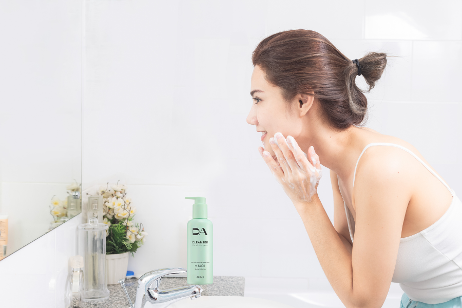 Người phụ nữ đang rửa mặt trong phòng tắm, bên cạnh là chai sữa rửa mặt màu xanh.