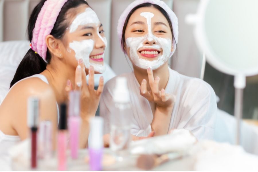 Hai người đang đắp mặt nạ dưỡng da, cười vui vẻ trước gương với nhiều sản phẩm chăm sóc da xung quanh.