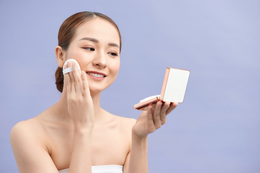 Người phụ nữ cầm gương trang điểm và dùng bông phấn, nền xanh nhạt.