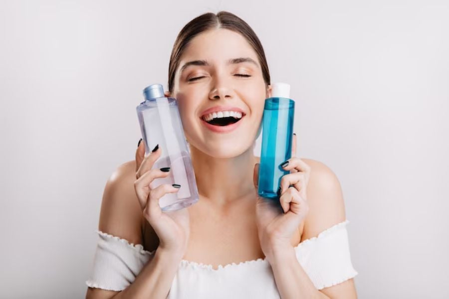 Người phụ nữ cười tươi, cầm hai chai mỹ phẩm màu xanh và tím nhạt, trên nền trắng.