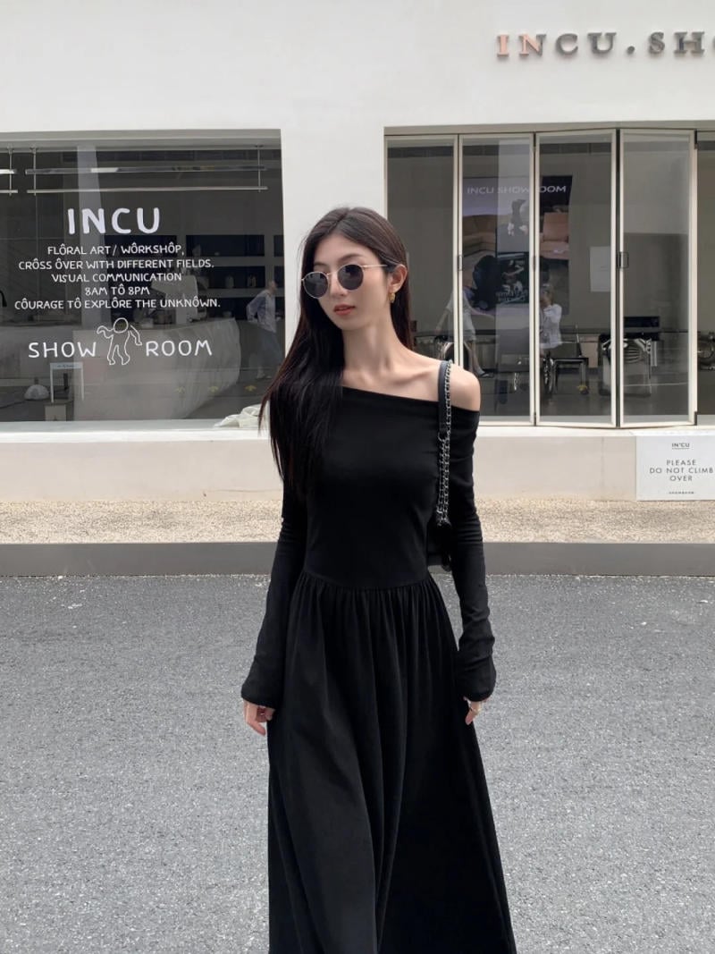 Người mặc váy đen dài đứng trước cửa hàng kính, đeo kính râm, tay cầm túi xách trong khung cảnh đường phố.