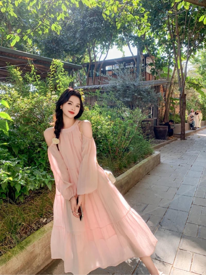 Người phụ nữ mặc váy hồng đứng trên lối đi lát đá, xung quanh là cây cối xanh tươi dưới ánh sáng mặt trời.