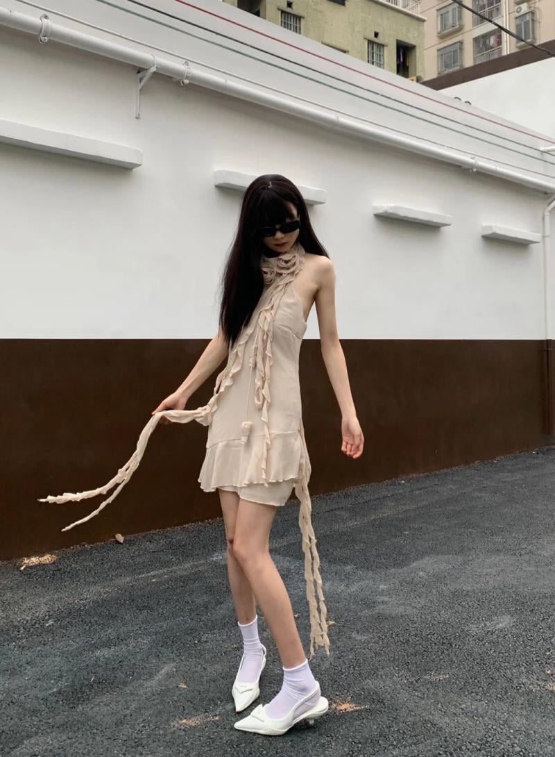 Người mặc váy màu kem với phụ kiện dài, đi giày trắng, đứng ngoài trời trước bức tường trắng và nâu.