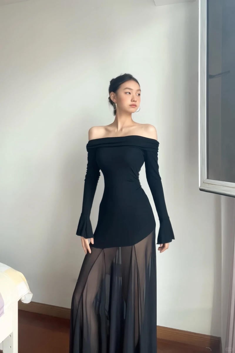 Người mẫu mặc váy đen trễ vai với phần dưới xuyên thấu, đứng cạnh cửa sổ trong phòng sáng.