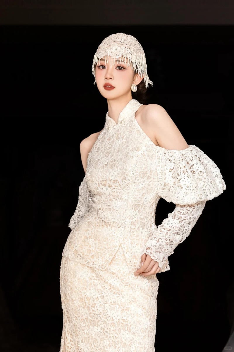 Người mẫu mặc váy ren trắng, đội mũ cùng tông, đứng trước nền đen.