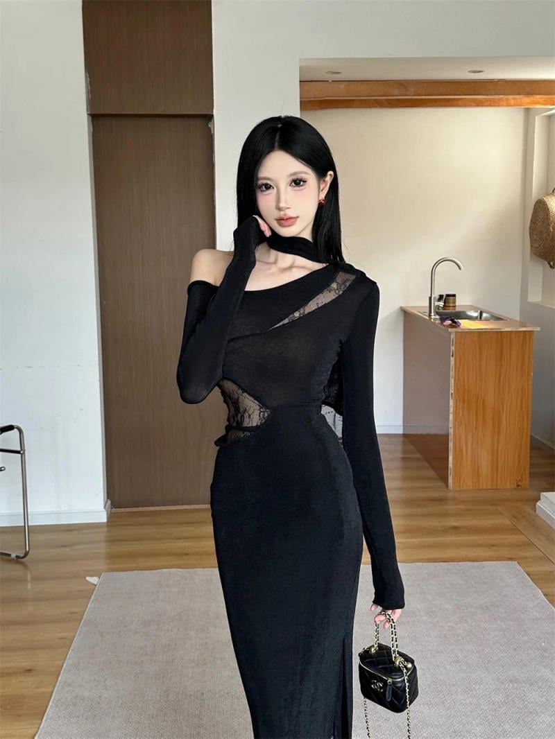 Người mặc váy đen dài đứng trong phòng, tay cầm túi xách nhỏ, nền là khu vực bếp gỗ.