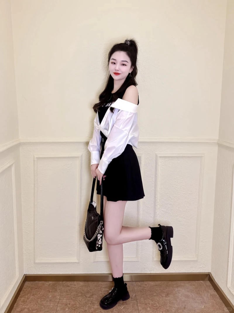 Người phụ nữ đứng dựa vào tường, mặc áo sơ mi trắng, váy đen, cầm túi xách, tạo dáng với chân hơi nhấc lên.