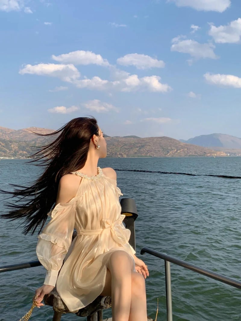 Người phụ nữ mặc váy ngồi bên bờ hồ, tóc bay trong gió, phía sau là núi non và bầu trời nhiều mây.
