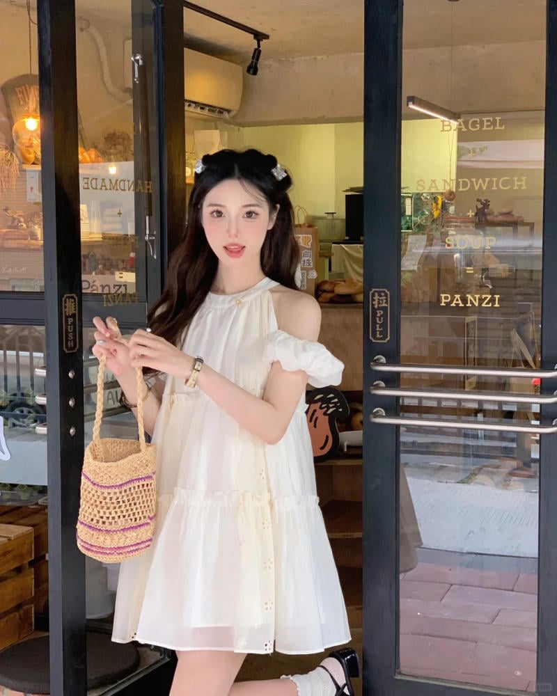 Người phụ nữ mặc váy trắng đứng trước cửa tiệm, tay cầm túi cói, phía sau là nội thất quán cà phê.