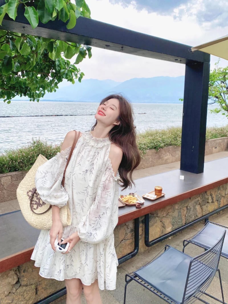 Người phụ nữ đứng cười trước biển, mặc váy trắng, cầm túi cói, trên bàn có đồ ăn và đồ uống.