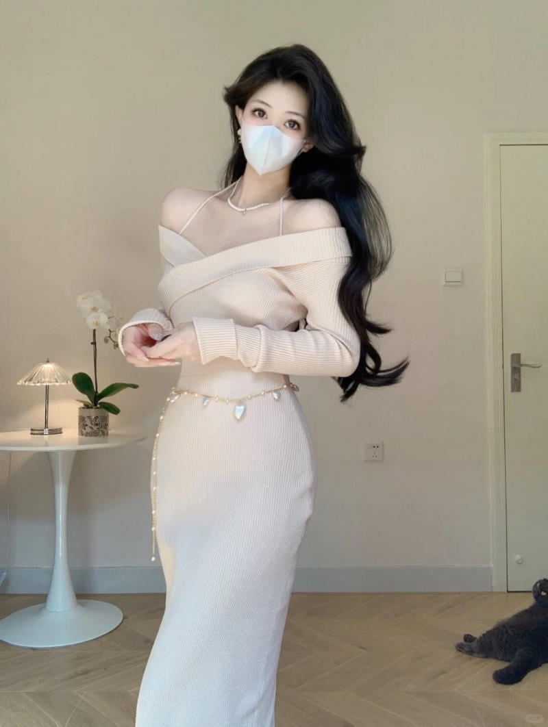 Người đeo khẩu trang, mặc váy dài màu sáng, đứng trong phòng với bàn trang trí và một chú mèo nằm dưới sàn.