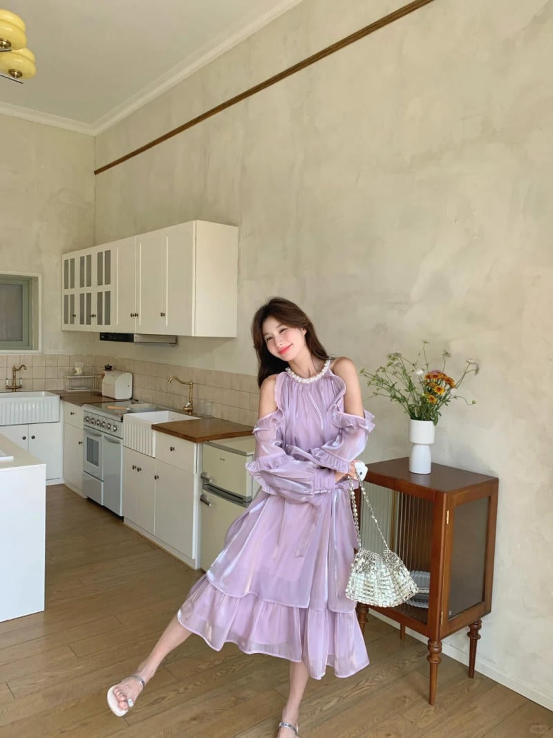 Người phụ nữ mặc váy tím đứng trong bếp sáng sủa, tay cầm túi xách, cạnh tủ kính và bình hoa.
