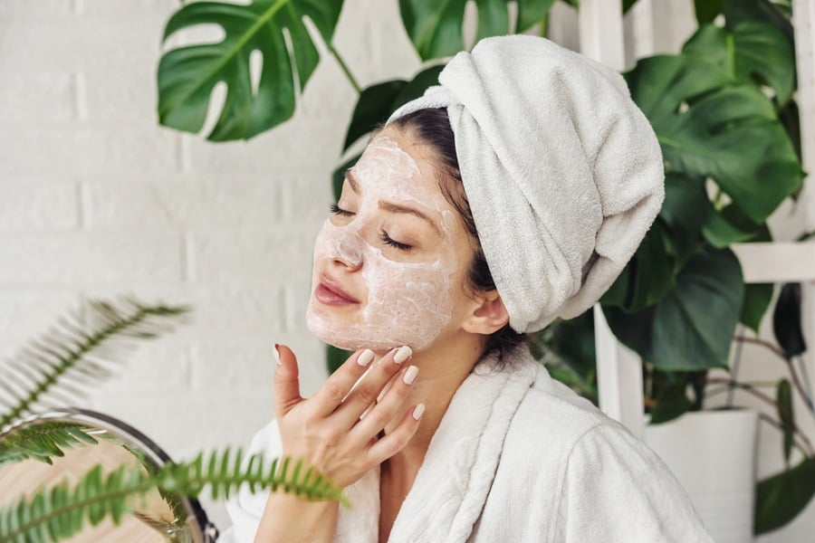 Người đang dưỡng da mặt với khăn tắm quấn trên đầu, nền là cây xanh và tường gạch trắng.