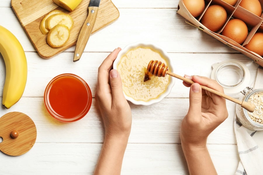Hai tay cầm bát hỗn hợp mật ong và lát chuối, trên bàn có trứng, yến mạch và mật ong.
