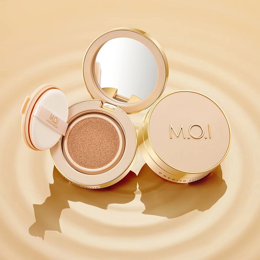 Hộp phấn cushion màu vàng với gương, bông phấn và nắp có chữ "M.O.I" trên nền nâu nhạt.