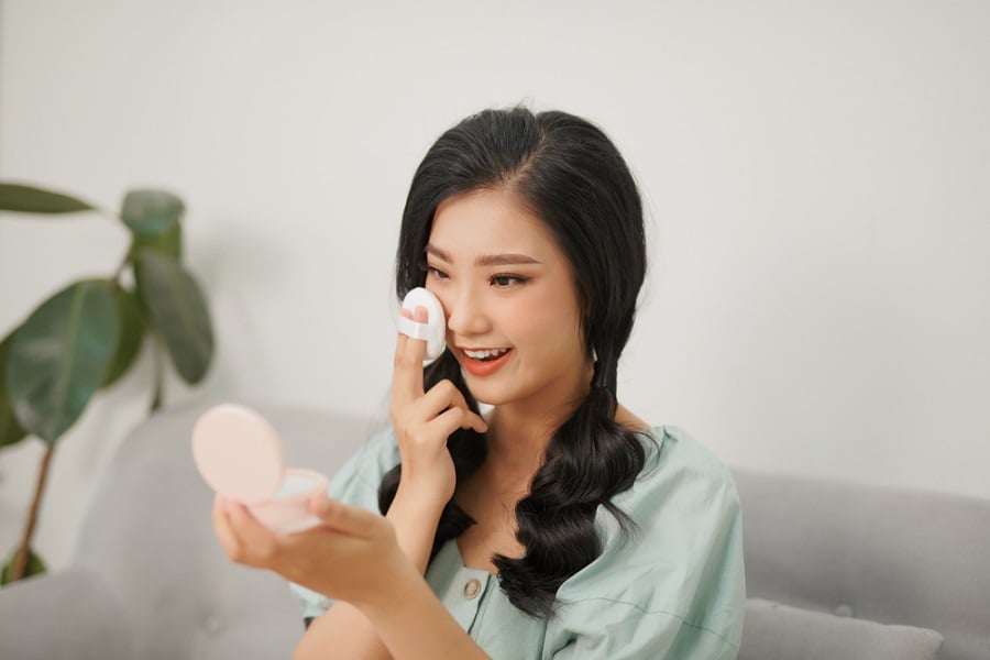 Người phụ nữ đang ngồi trên ghế, trang điểm bằng phấn từ hộp trang điểm, với cây xanh phía sau.