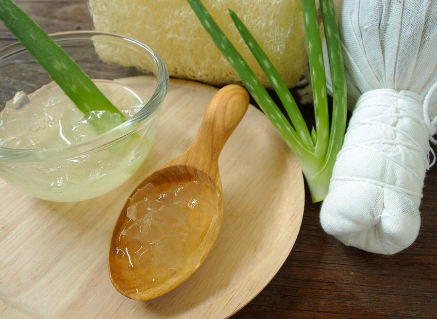 Lá nha đam và gel trong bát thủy tinh và muỗng gỗ trên bàn gỗ, bên cạnh là một chiếc khăn trắng cuộn tròn.