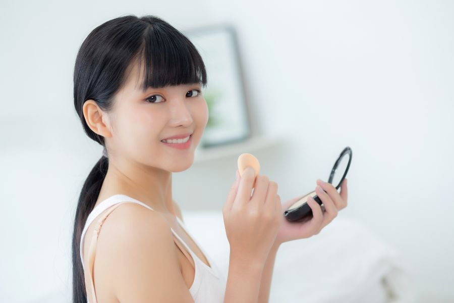 Người phụ nữ cầm gương và bông phấn, mỉm cười trong không gian sáng sủa.