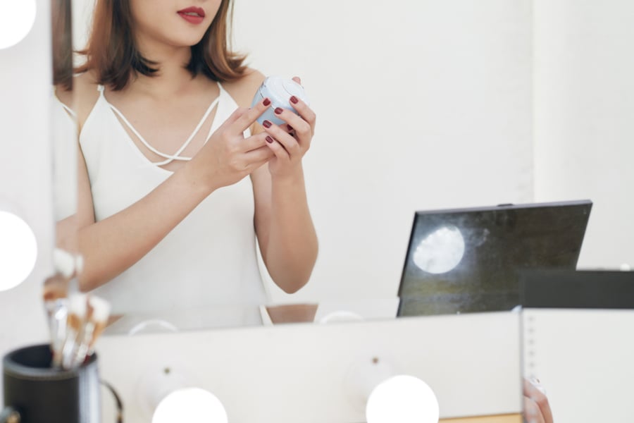Người phụ nữ đang cầm hộp mỹ phẩm trước gương trang điểm, bên cạnh là máy tính xách tay.