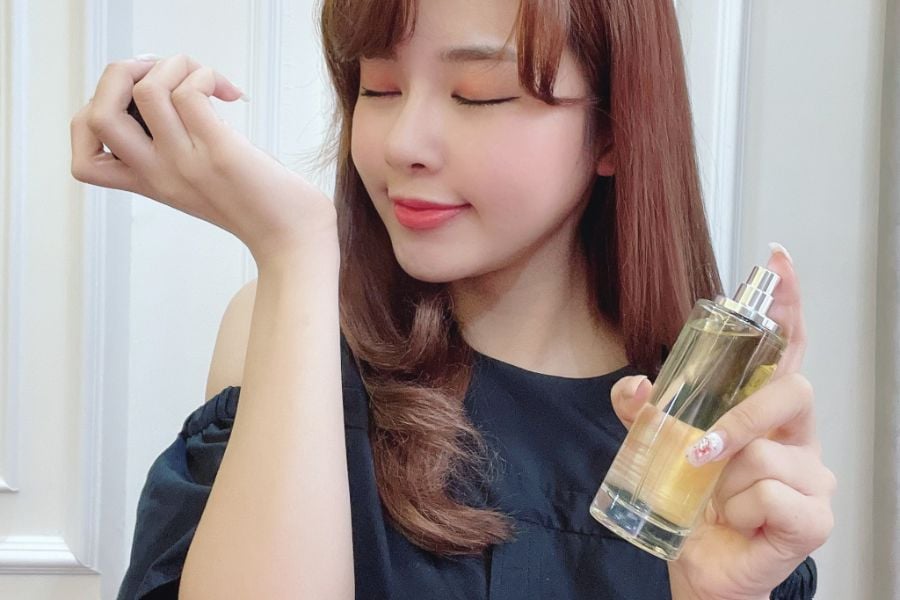 Người phụ nữ cầm chai nước hoa, nhắm mắt và mỉm cười, mặc áo đen, tóc dài màu nâu.