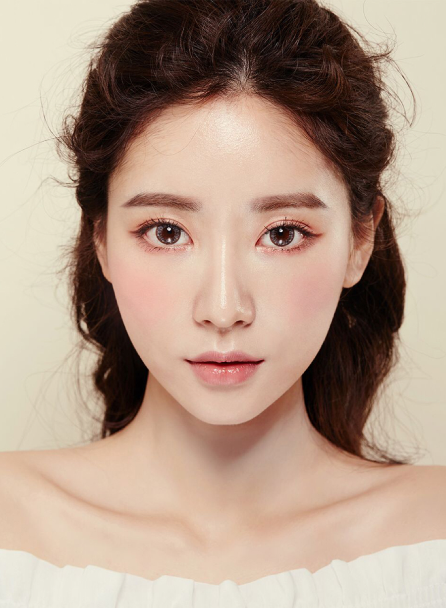 Người phụ nữ với mái tóc đen dài, trang điểm nhẹ nhàng, gương mặt nhìn thẳng vào máy ảnh, nền màu kem.