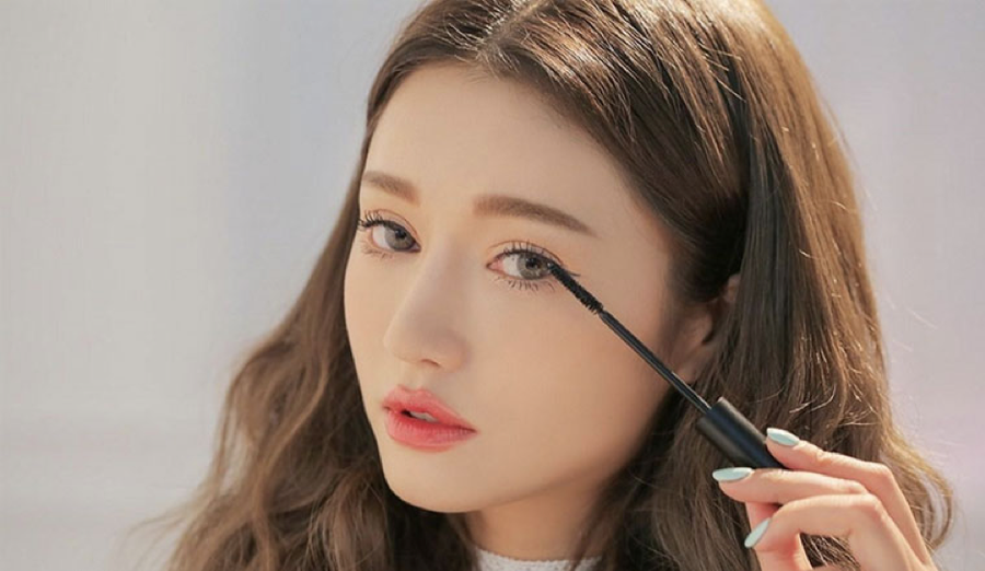 Người phụ nữ đang chuốt mascara lên lông mi, tóc dài xoăn nhẹ, ánh sáng tự nhiên chiếu vào khuôn mặt.