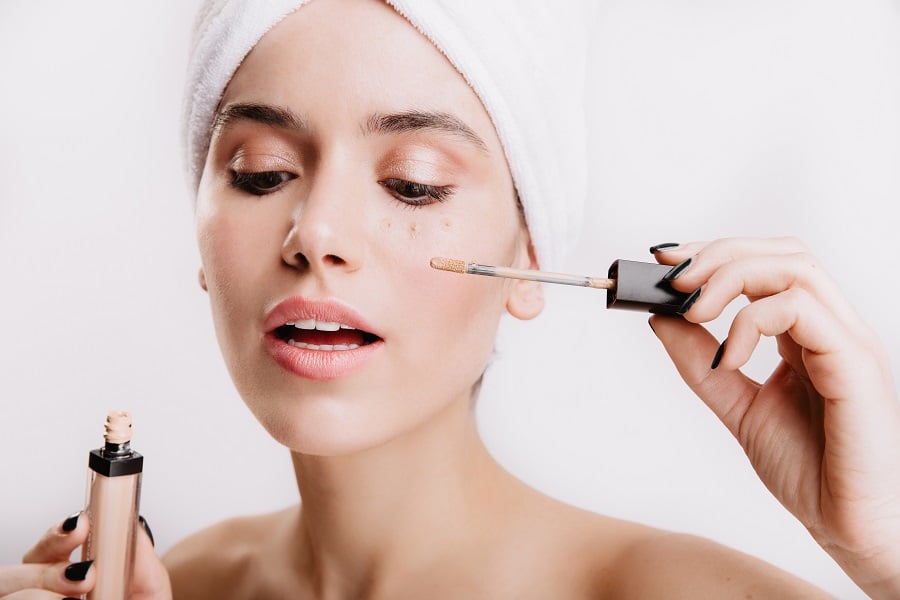 Người đang thoa kem che khuyết điểm lên mặt, đầu quấn khăn trắng, nền trắng.