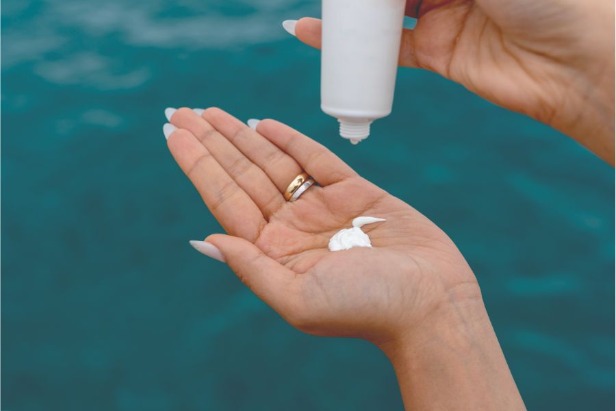 Bàn tay đang bóp kem từ chai nhỏ màu trắng, với nền biển xanh phía sau.