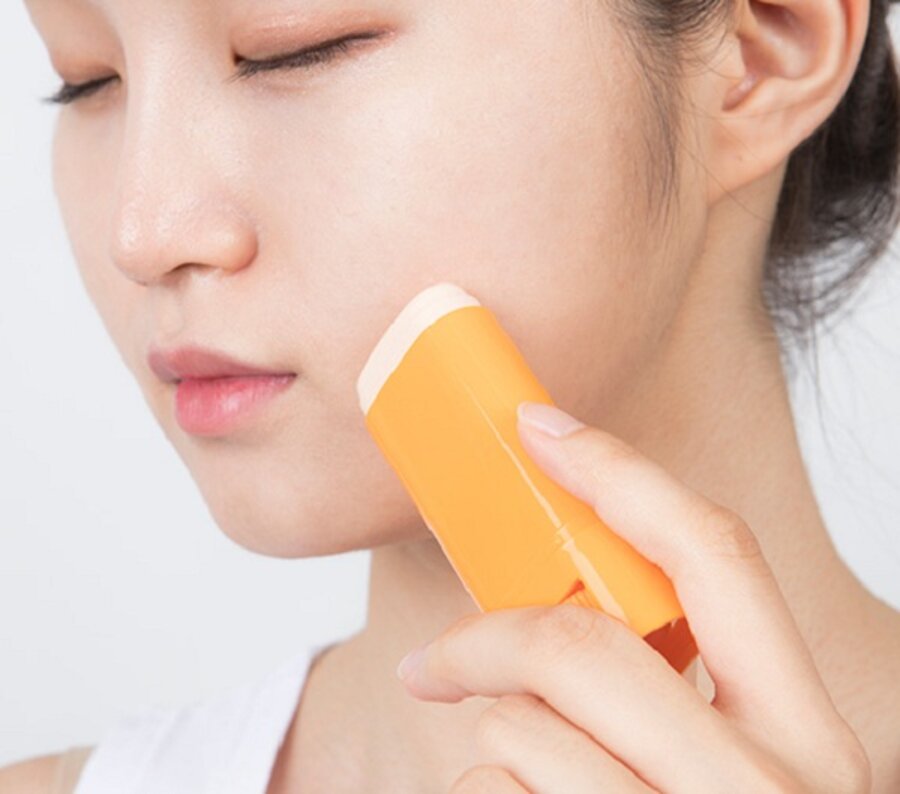 Người đang sử dụng thanh lăn màu cam trên da mặt, mắt nhắm, mặc áo trắng.