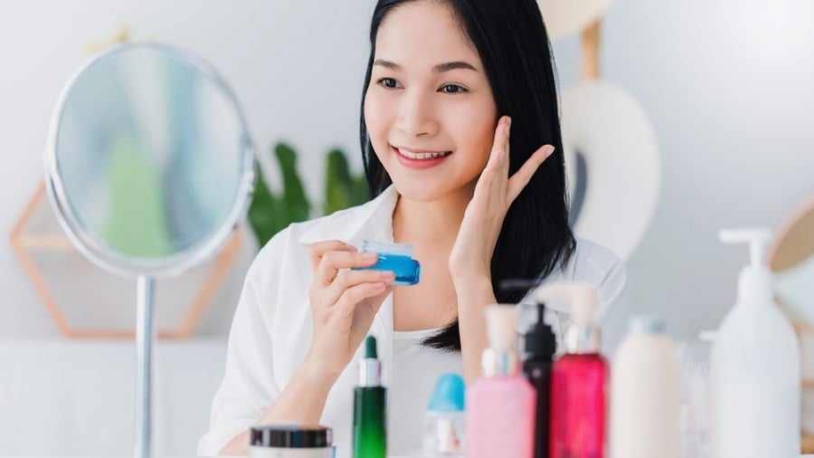 Người phụ nữ cầm lọ kem, ngồi trước gương, xung quanh là các sản phẩm chăm sóc da.