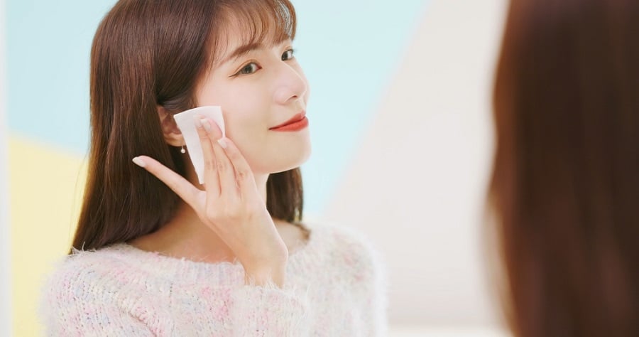 Người phụ nữ đứng trước gương, sử dụng bông tẩy trang lau mặt, nền màu pastel.