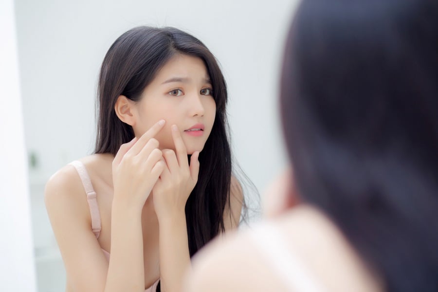 Người phụ nữ đứng trước gương, tay chạm vào mặt, có vẻ đang quan sát da mặt của mình.