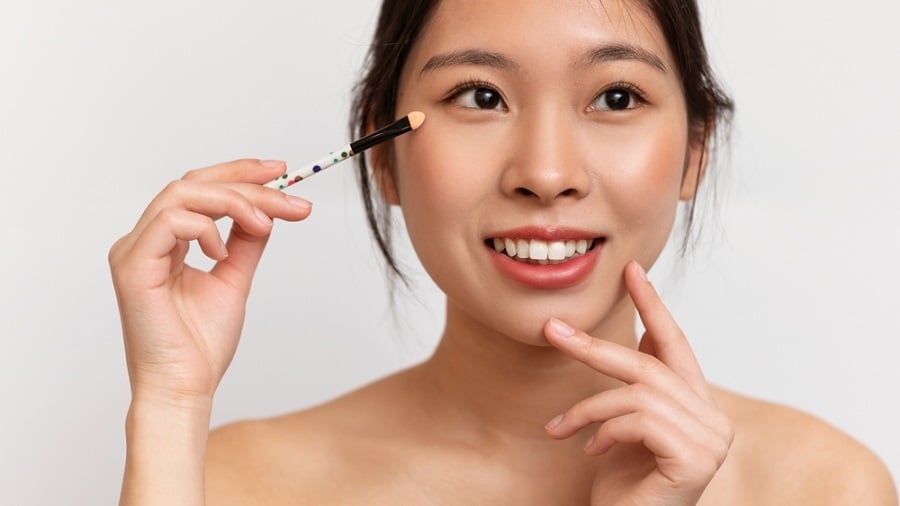 Người cầm cọ trang điểm gần mắt, mỉm cười, nền trắng.