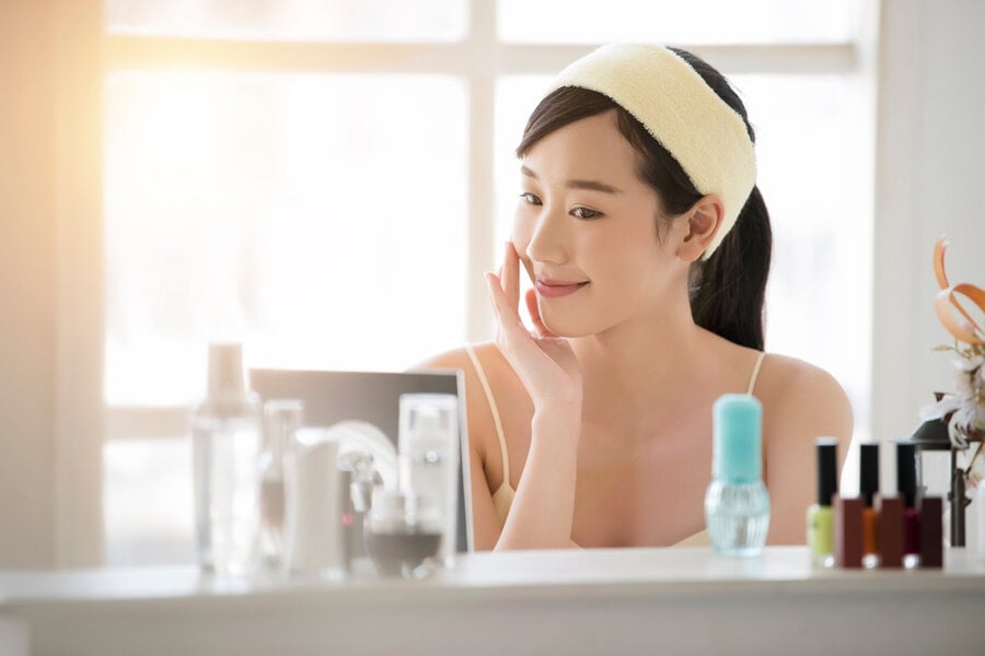 Người phụ nữ ngồi trước gương, đội băng đô, chạm vào má, trên bàn có nhiều sản phẩm chăm sóc da và lọ sơn móng tay.