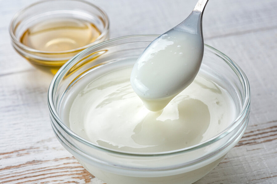 Một bát sữa chua trắng với một muỗng được nhấc lên, phía sau có một bát nhỏ đựng chất lỏng màu vàng.