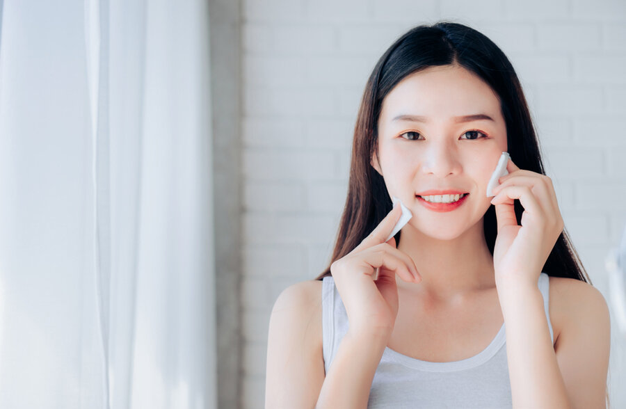Người phụ nữ đang mỉm cười, dùng bông tẩy trang trên mặt trong không gian sáng sủa, nền gạch trắng.