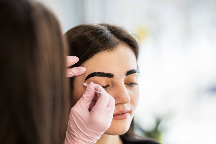Người thợ đang vẽ lông mày cho một người, sử dụng găng tay và bút chuyên dụng trong tiệm làm đẹp.