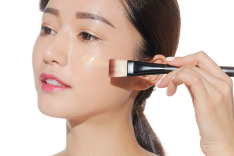 Người cầm cọ trang điểm, thoa kem nền lên má, nền trắng, thể hiện quá trình trang điểm.