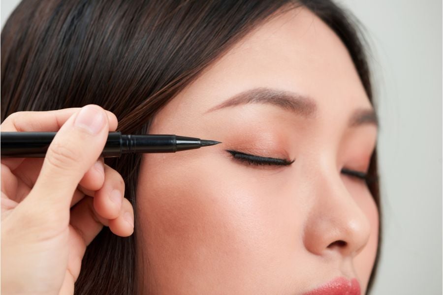 Người đang được kẻ mắt với bút kẻ mắt đen, mắt nhắm và môi hồng nhạt, tay cầm bút ở phía bên trái.