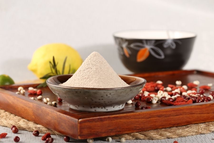 Bát bột trắng trên khay gỗ cùng hạt, trái cây và bát khác ở nền phía sau.