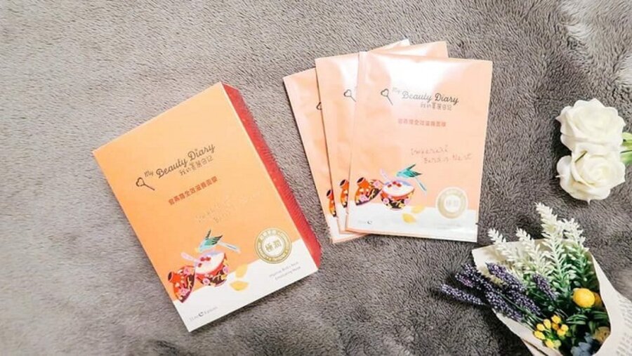 Hộp và gói mặt nạ dưỡng da đặt trên thảm xám, bên cạnh là hoa trắng và tím.