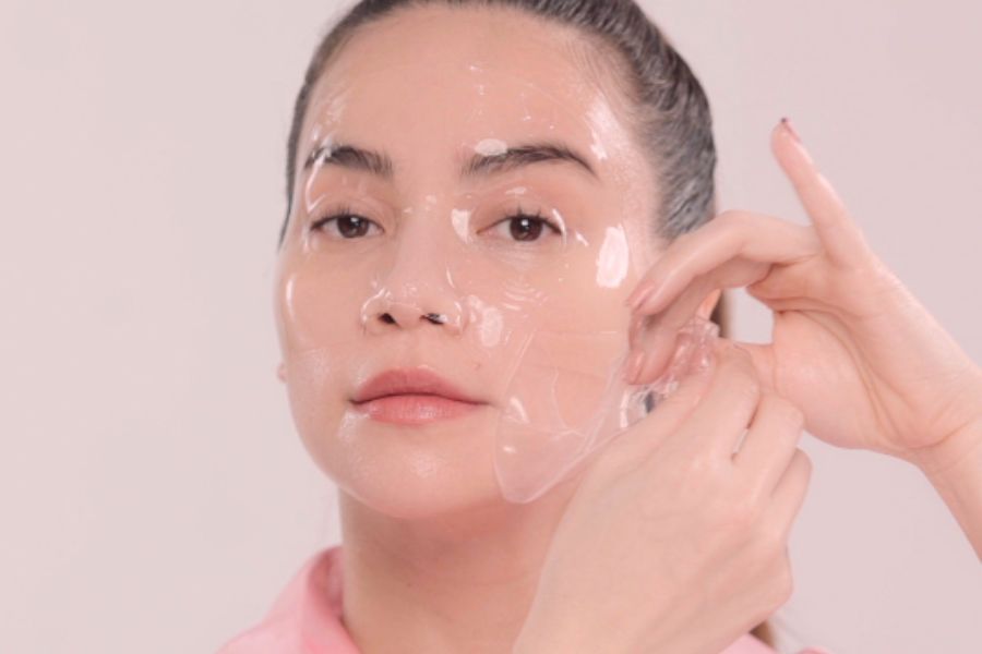 Người đang tháo mặt nạ dưỡng da trong suốt khỏi mặt, nền màu hồng nhạt.