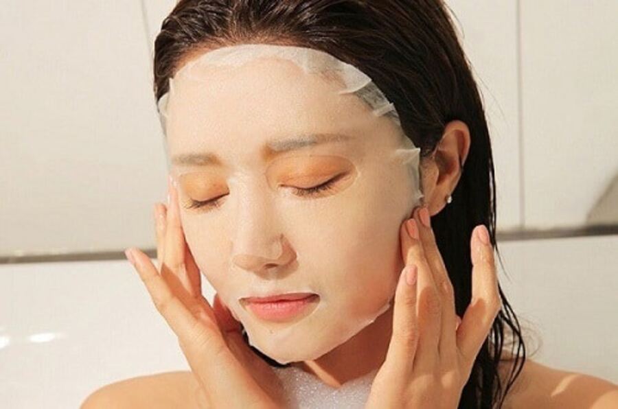 Người đang đắp mặt nạ dưỡng da, mắt nhắm, tay chạm nhẹ vào mặt, tóc ướt, trong không gian sáng.