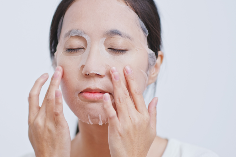 Người phụ nữ đắp mặt nạ dưỡng da trên mặt, nhắm mắt và chạm nhẹ vào má, nền trắng.