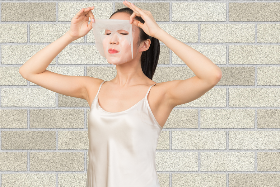 Người phụ nữ trong trang phục trắng đang đắp mặt nạ dưỡng da, đứng trước bức tường gạch sáng màu.