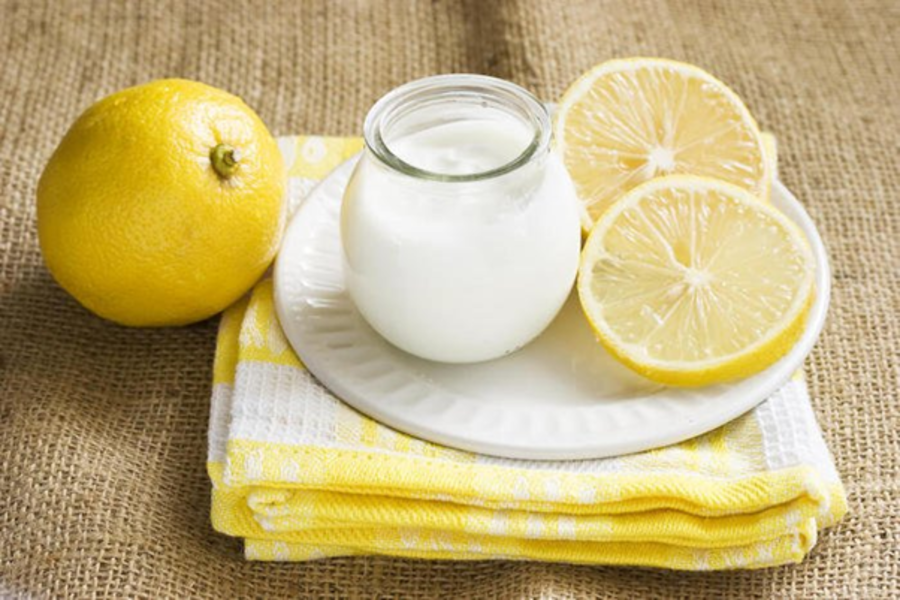 Hũ sữa chua trên đĩa trắng, bên cạnh là chanh vàng nguyên trái và cắt lát, đặt trên khăn màu vàng.