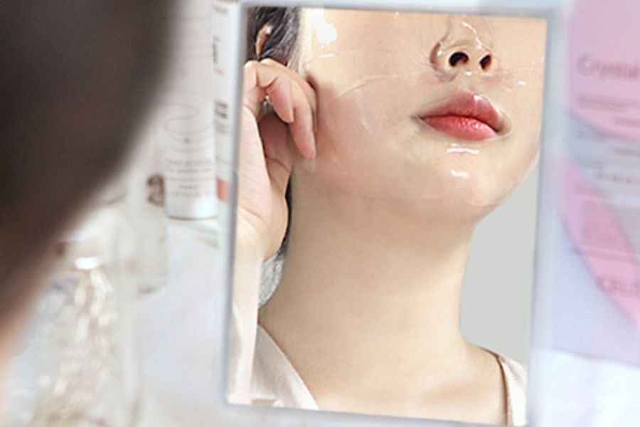 Người đang nhìn vào gương với mặt nạ dưỡng da trong suốt, môi hồng, ánh sáng nhẹ từ bên trái.