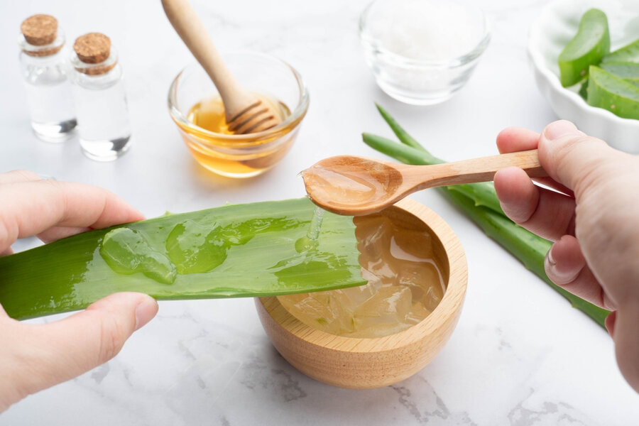 Người đang cạo gel từ lá nha đam vào bát gỗ, bên cạnh là mật ong và các nguyên liệu khác trên bàn.